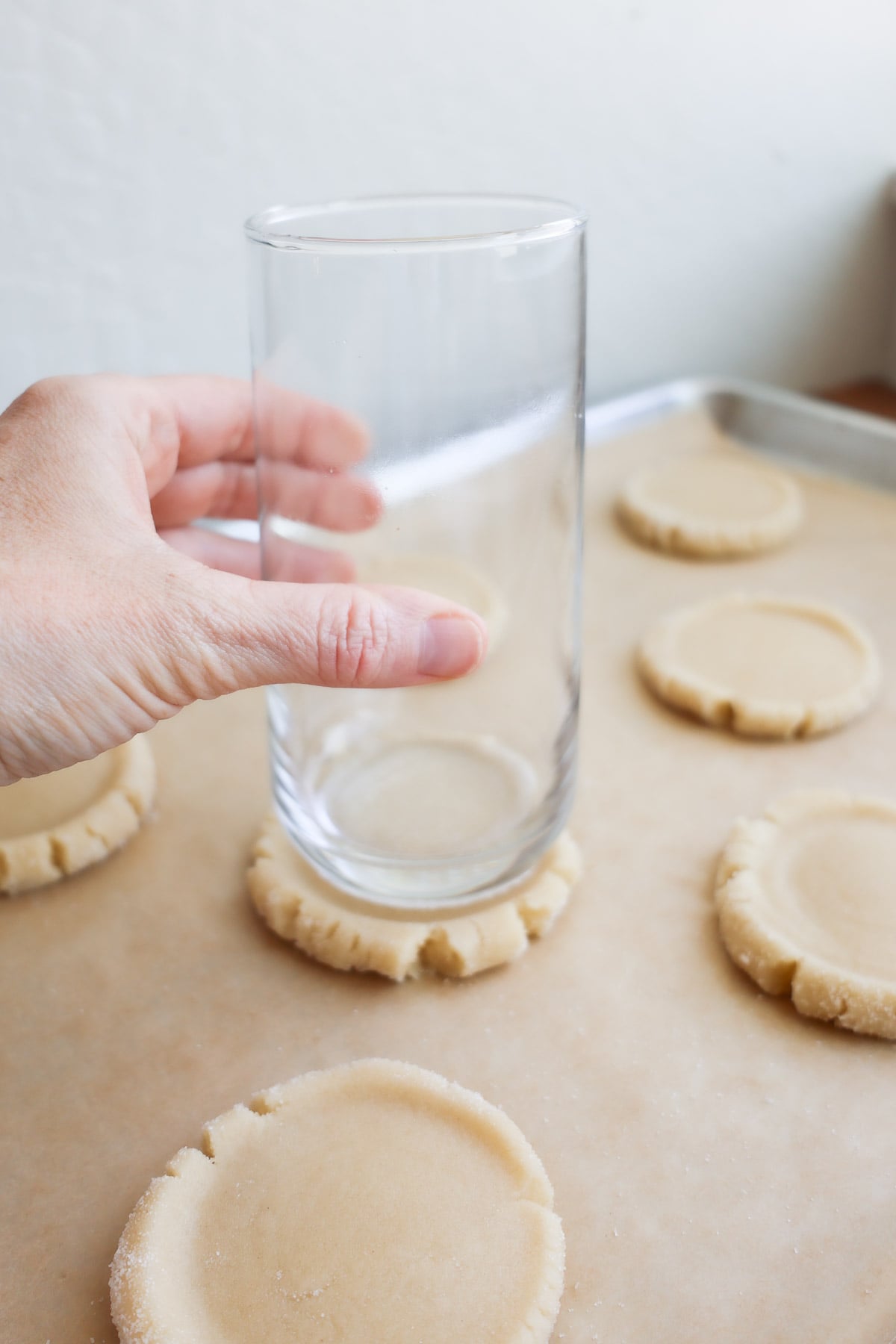 A hand holding a glass, flattening a sugar cookie with the bottom, on a parchment lined baking sheet.
