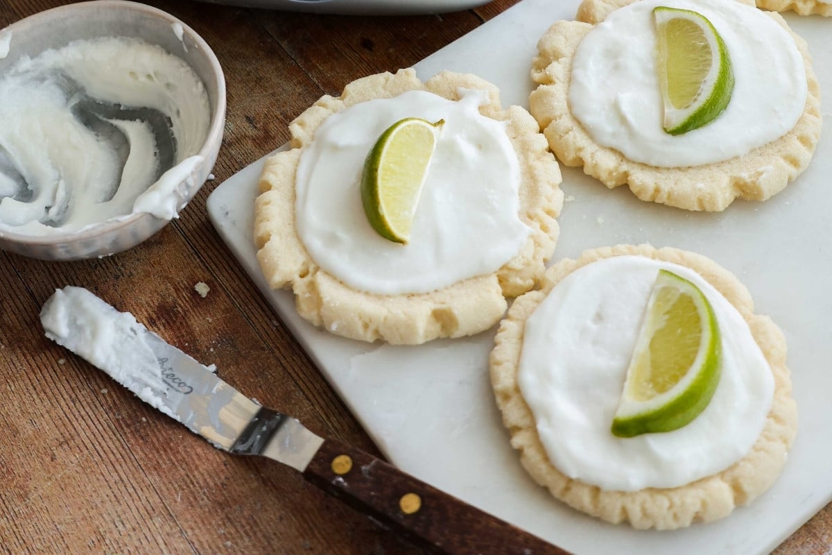 Coconut lime cookies on a marble cutting board, with a bowl of icing and an offset spatula.