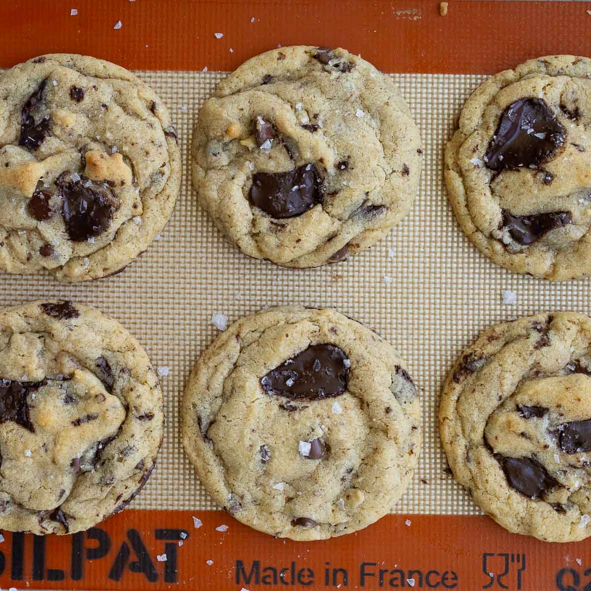 Baked, brown butter sourdough chocolate chip cookies with salt flakes on top, on a baking sheet lined with a silicone baking mat.