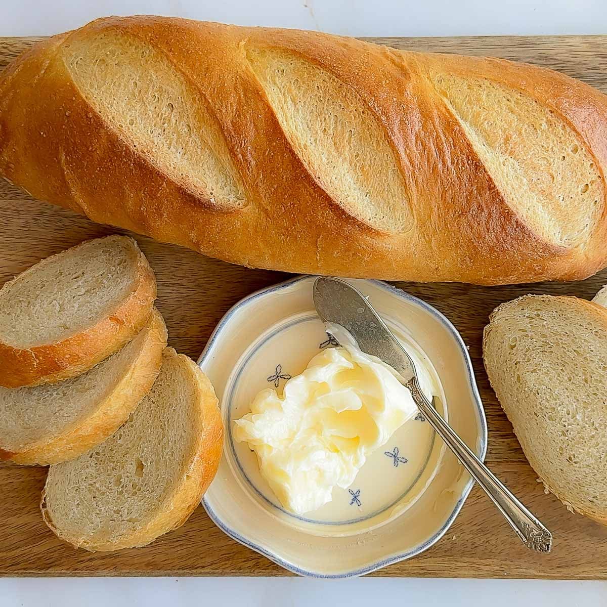A loaf of French bread on a wood cutting board, with sliced French bread and butter.
