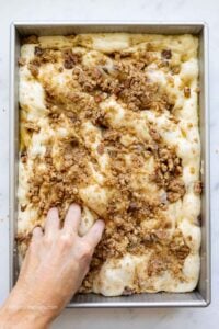 Chocolate chip banana bread focaccia with brown sugar pecan crumble topping on top, and a hand dimpling the dough.