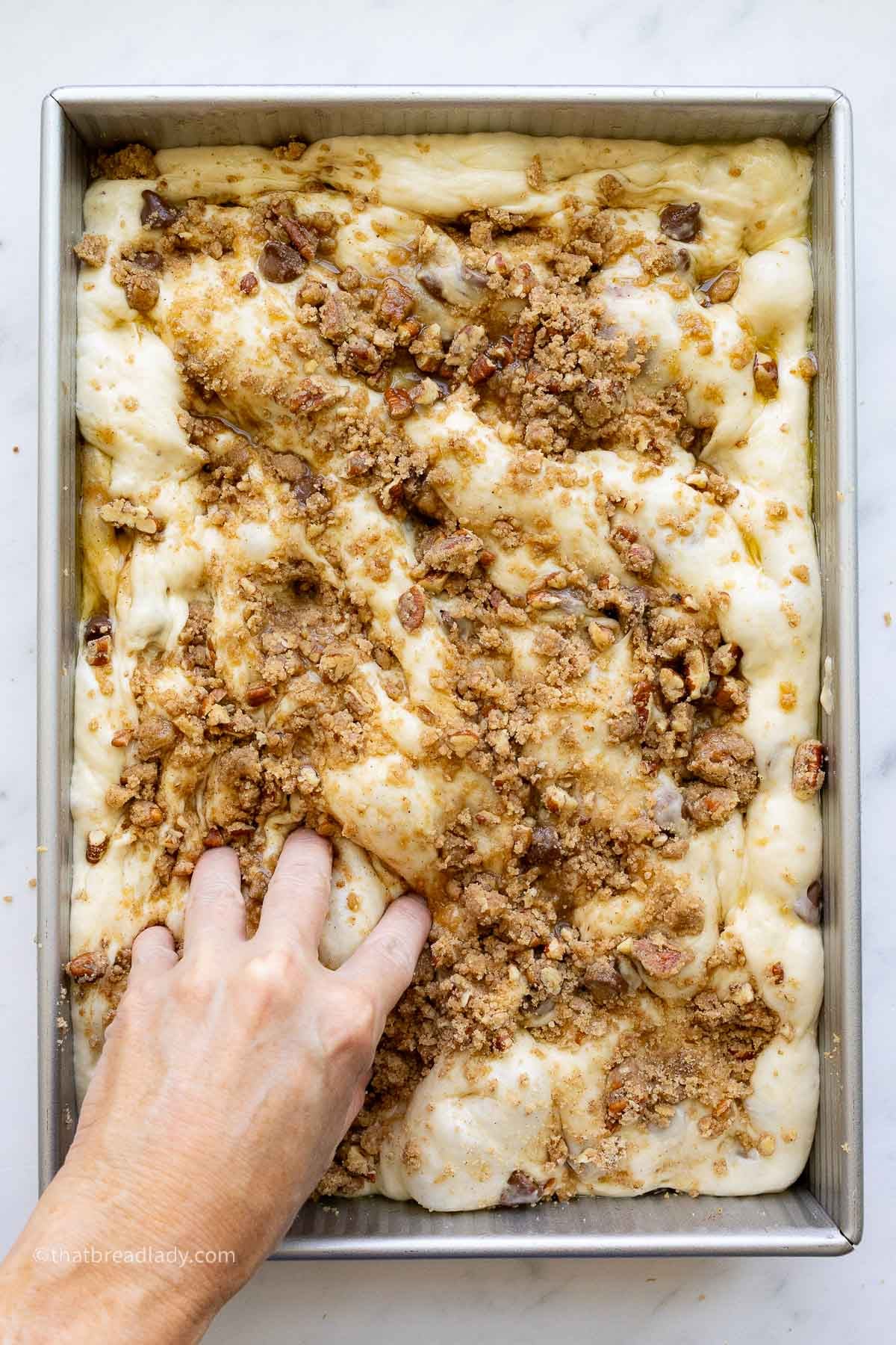 Chocolate chip banana bread focaccia dough in a 9x13-inch metal cake pan, the dough risen, with a brown sugar pecan crumble on top, and a hand dimpling the dough.