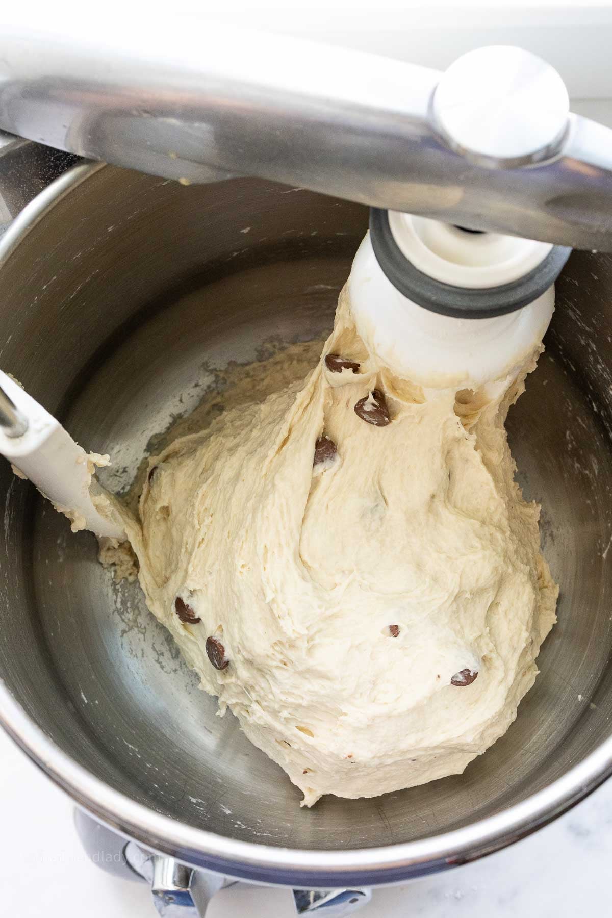 Chocolate chip banana bread focaccia dough in a stainless steel mixing bowl, with a dough roller attachment and bowl scraper attachment mixing the dough.