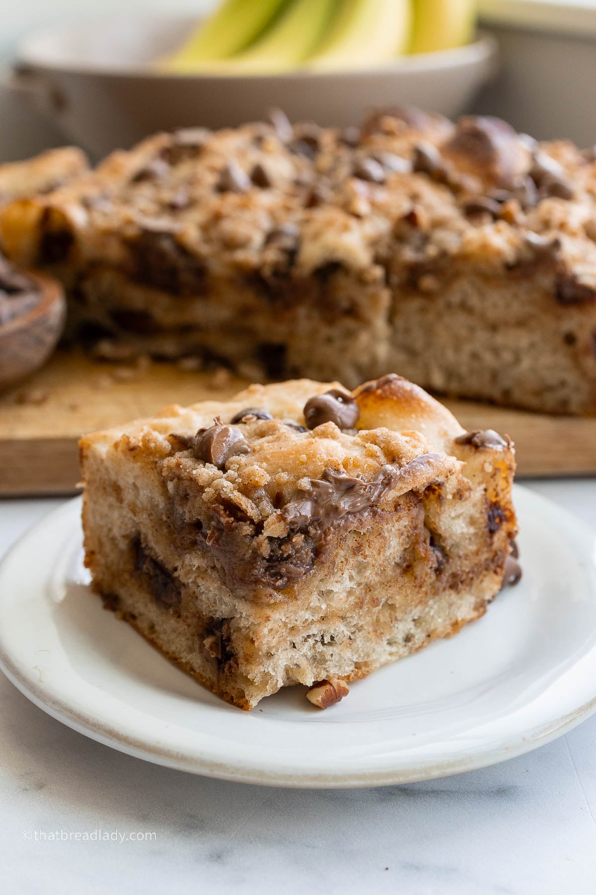A slice of chocolate chip banana bread focaccia on a plate, with more on the counter, resting on a wood cutting board.