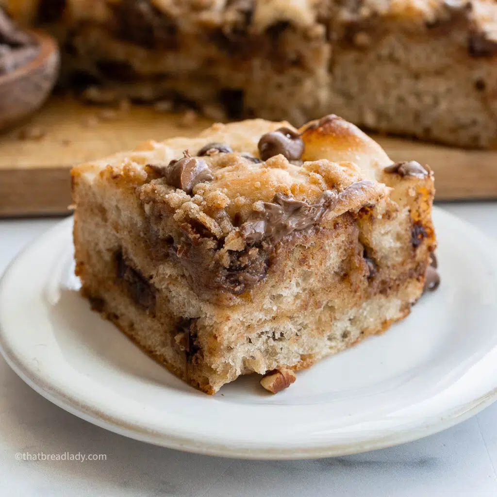 A slice of chocolate chip banana bread focaccia on a plate, with more focaccia behind it on a wood cutting board.