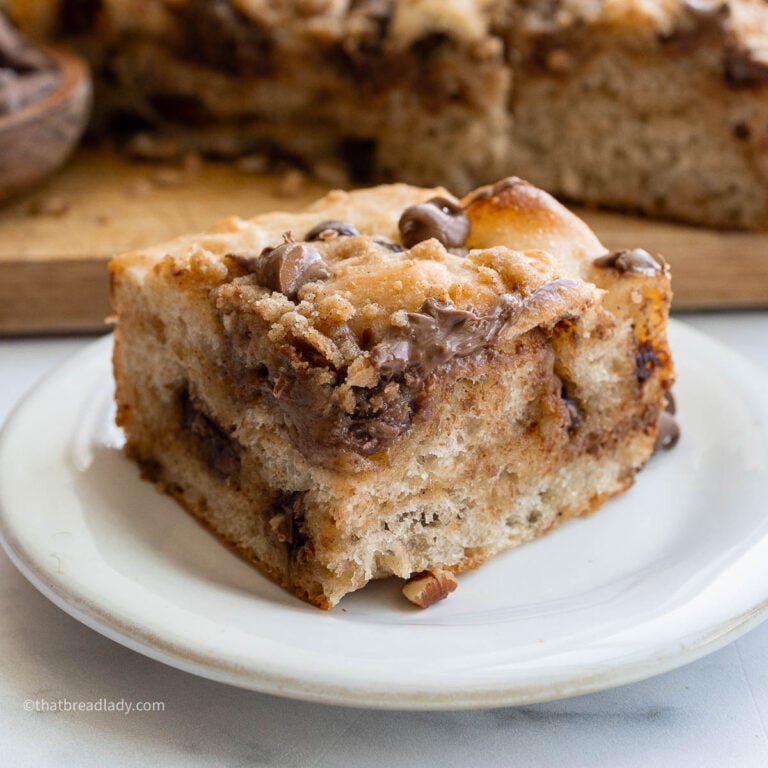 A slice of chocolate chip banana bread focaccia on a plate, with more focaccia behind it on a wood cutting board.