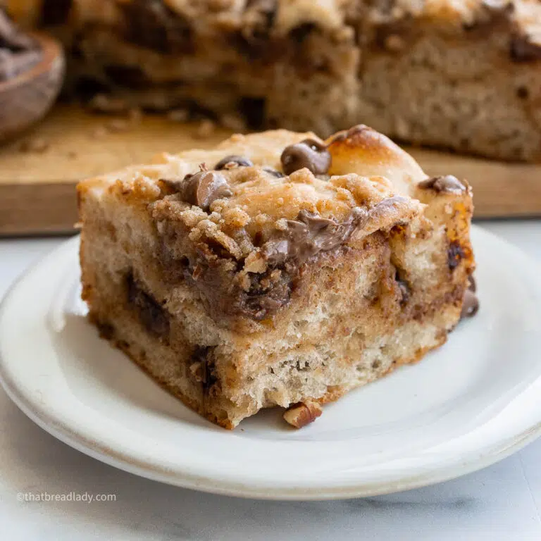 A slice of chocolate chip banana bread focaccia on a plate, with more focaccia behind it on a wood cutting board.