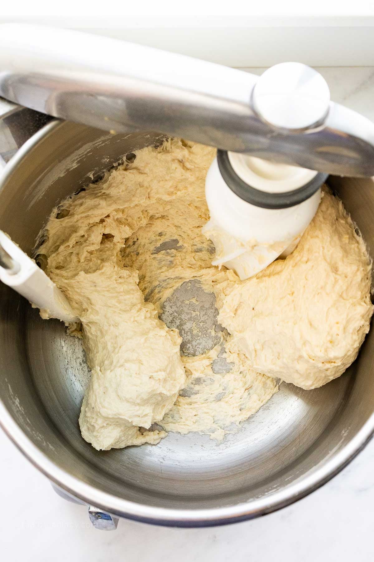 Bread dough in a stainless steel mixing bowl, with a dough roller attachment and bowl scraper attachment mixing the dough.