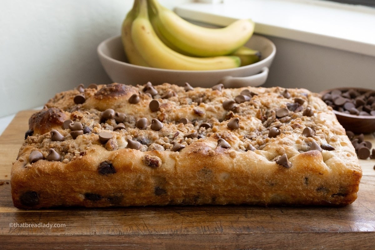 Chocolate chip banana bread focaccia on a wood cutting board with bananas and chocolate chips on the counter.