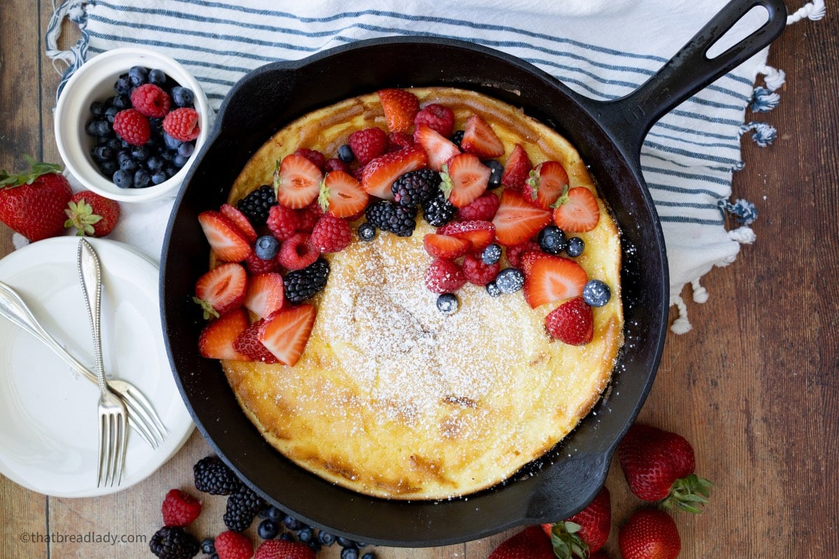 A baked sourdough dutch baby pancake, in a cast iron pan, topped with powdered sugar, strawberries, blueberries, and blackberries.