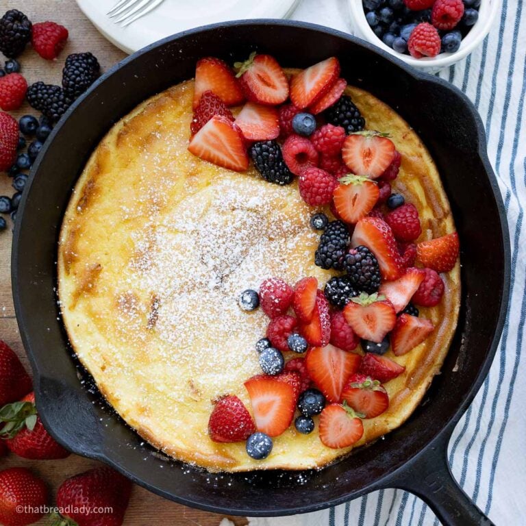 A cast iron skillet with a baked Dutch baby, topped with powdered sugar, strawberries, blackberries, and blueberries.