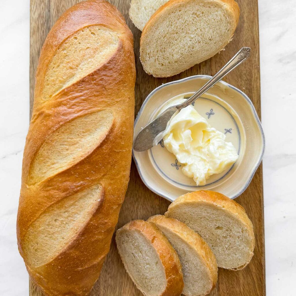 A loaf of french bread, sliced french bread, a butter dish, on a wood cutting board.
