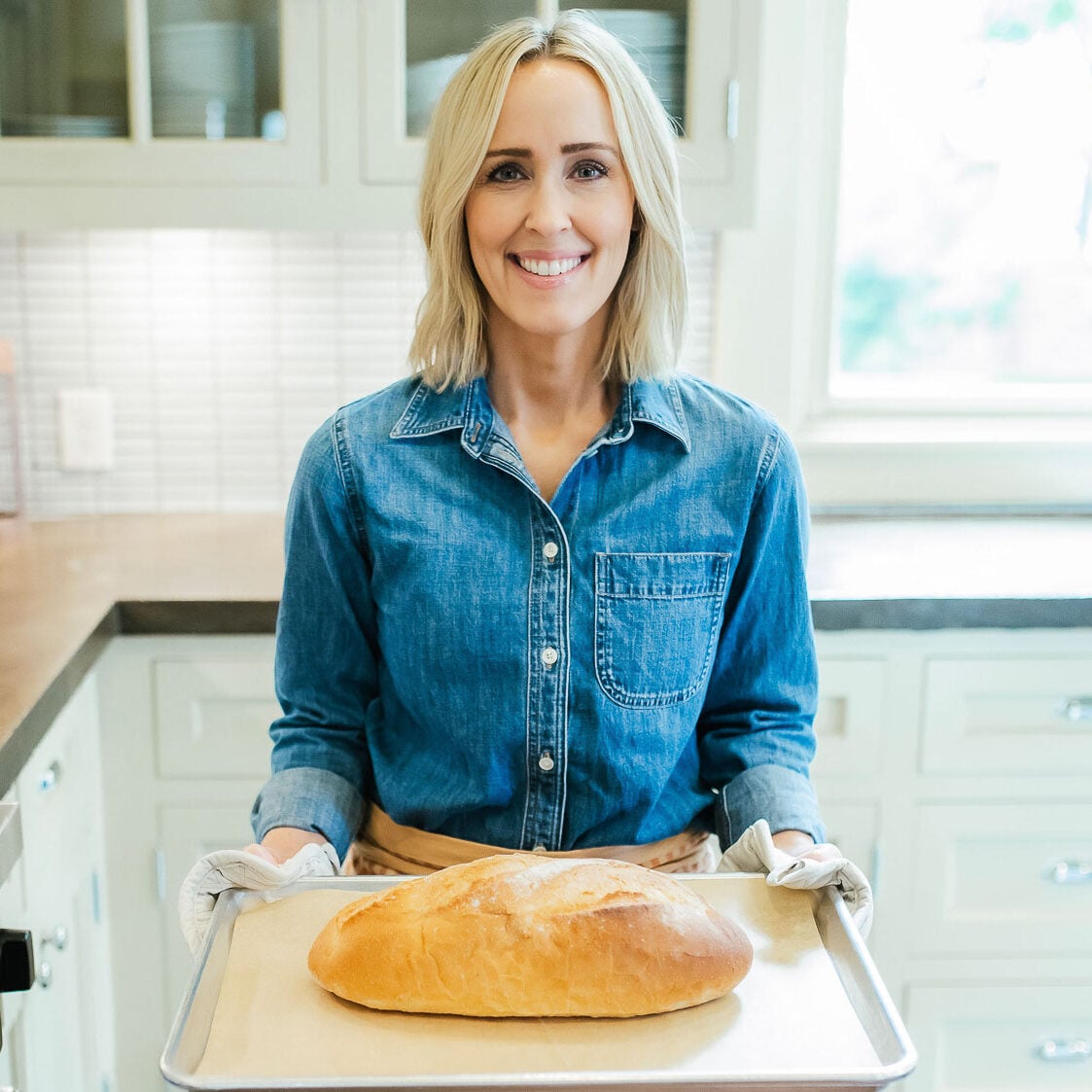 Heather Thomas, creator of That Bread Lady, standing in a kitchen, holding a baking sheet with a loaf of bread on it.