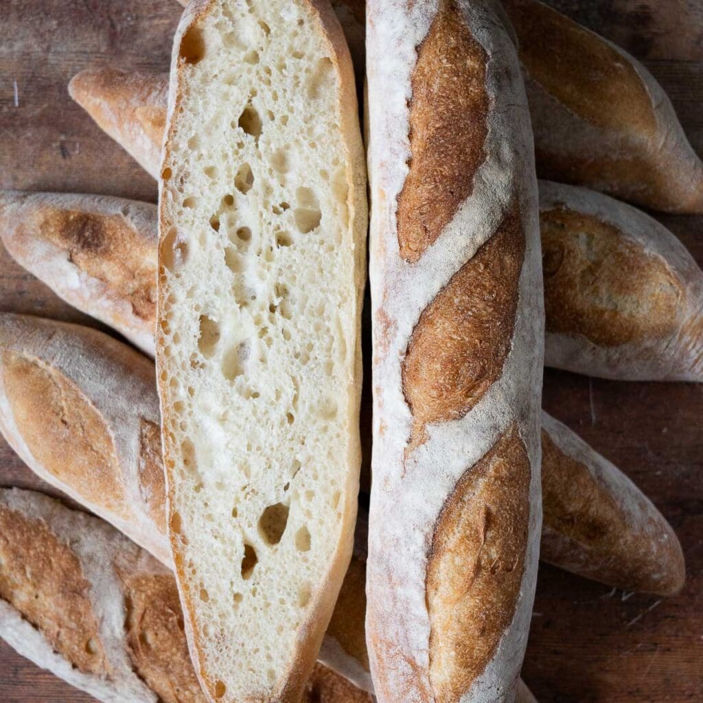 A pile of baguettes with two on top of the pile, one is sliced open lengthwise.