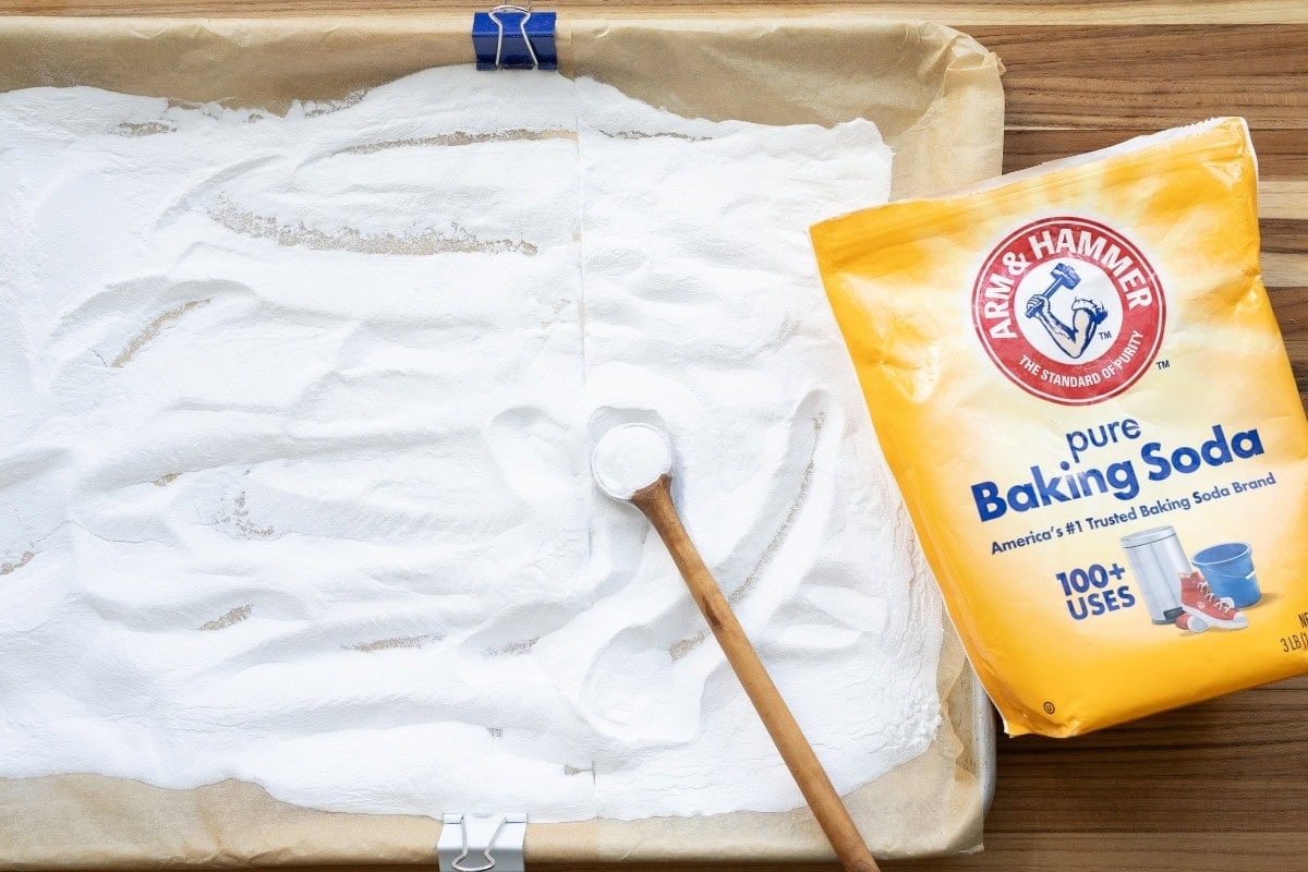 A baking sheet lined with parchment and filled with baking soda, with a wooden spoon resting in the baking soda, and a bag of baking soda resting on the wood board next to it.