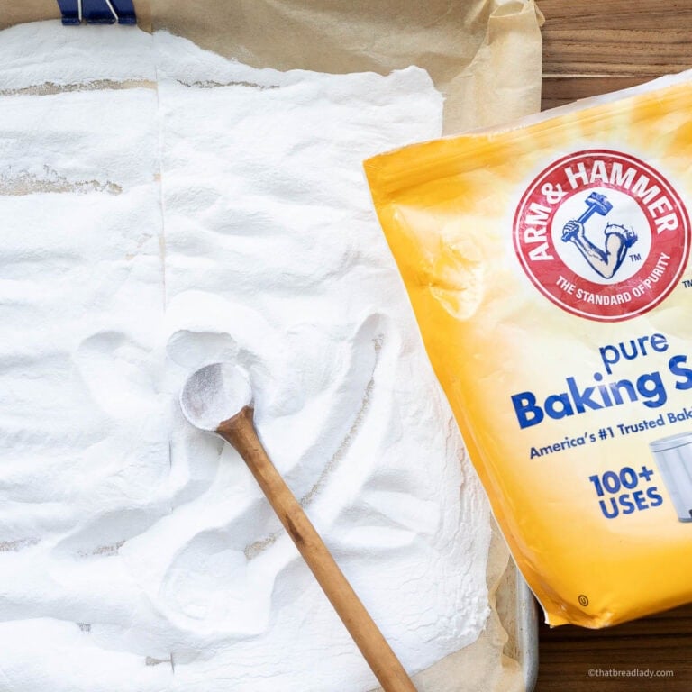 A baking sheet lined with parchment and filled with baking soda, with a wooden spoon resting in the baking soda, and a bag of baking soda resting on the wood board next to it.
