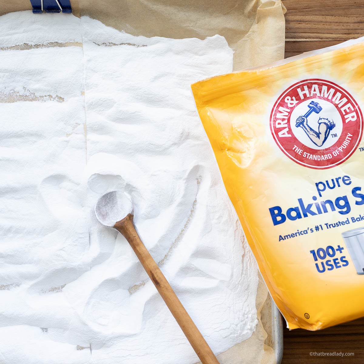 A baking sheet lined with parchment and filled with baking soda, with a wooden spoon resting in the baking soda, and a bag of baking soda resting on the wood board next to it.