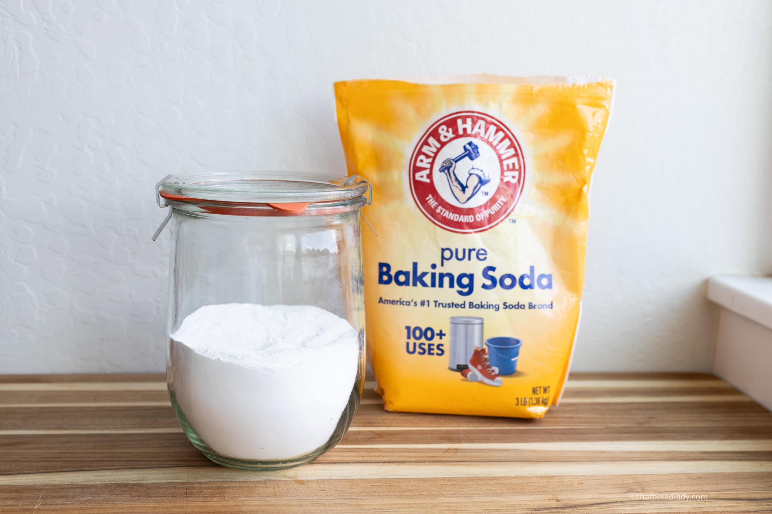 A glass jar of baked baking soda and a bag of baking soda, resting on a wood board.