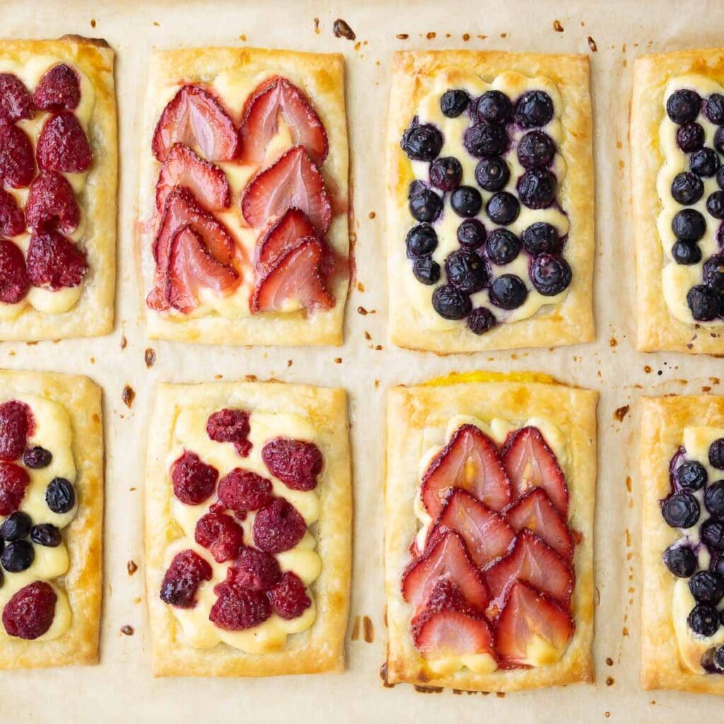 Several cream cheese puff pastries, with sliced strawberries, blueberries, and raspberries, on a baking sheet lined with parchment.