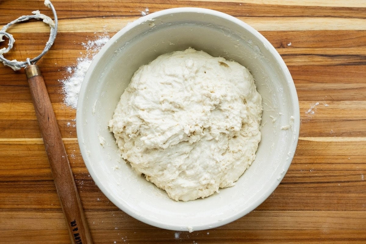 Mixed dough, in a mixing bowl, on a wood board with a Danish whisk
