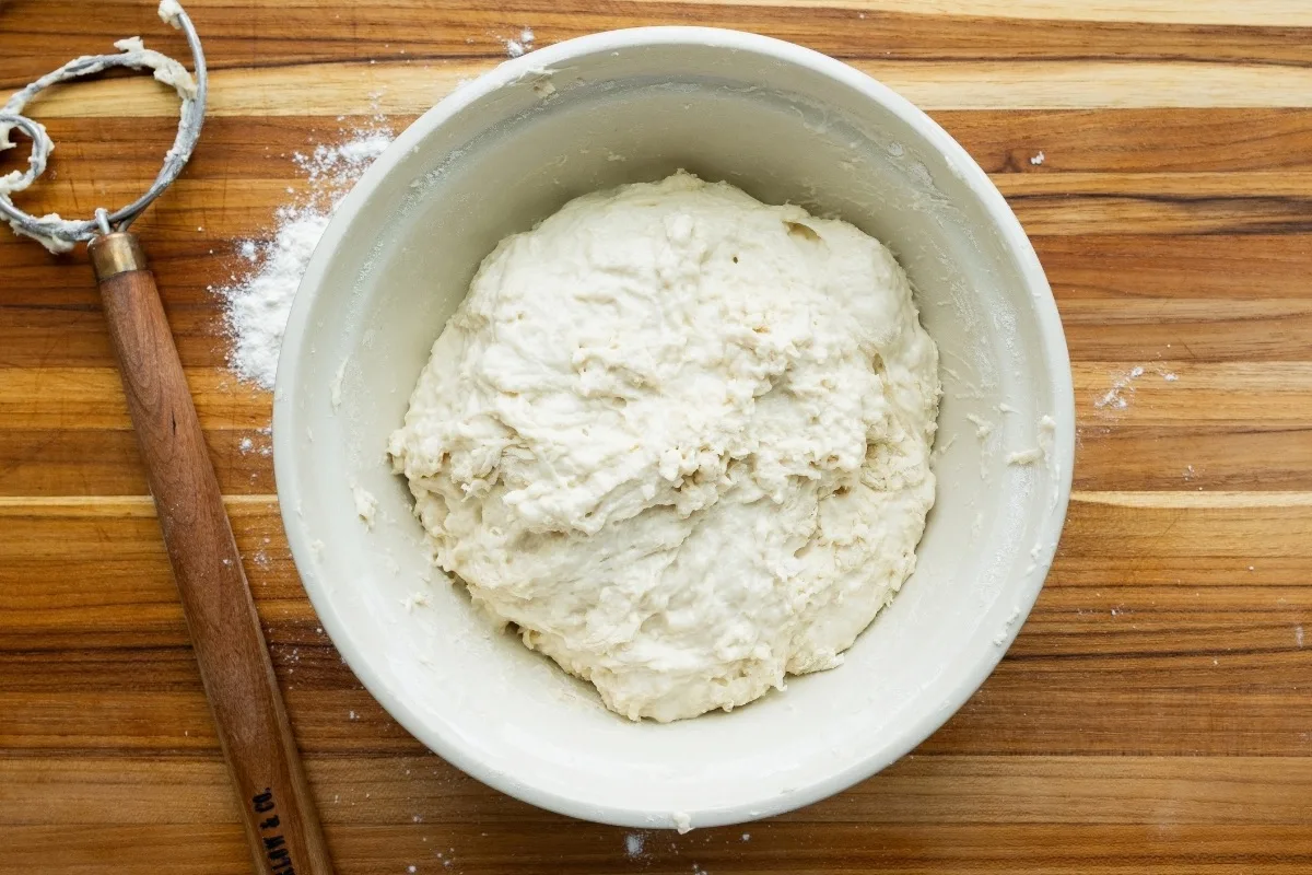 Mixed dough, in a mixing bowl, on a wood board with a Danish whisk