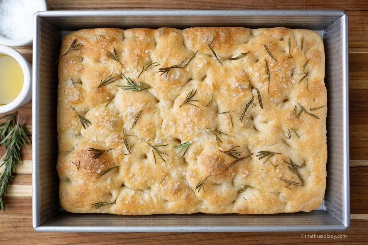 A pan of baked focaccia with rosemary leaves on top.