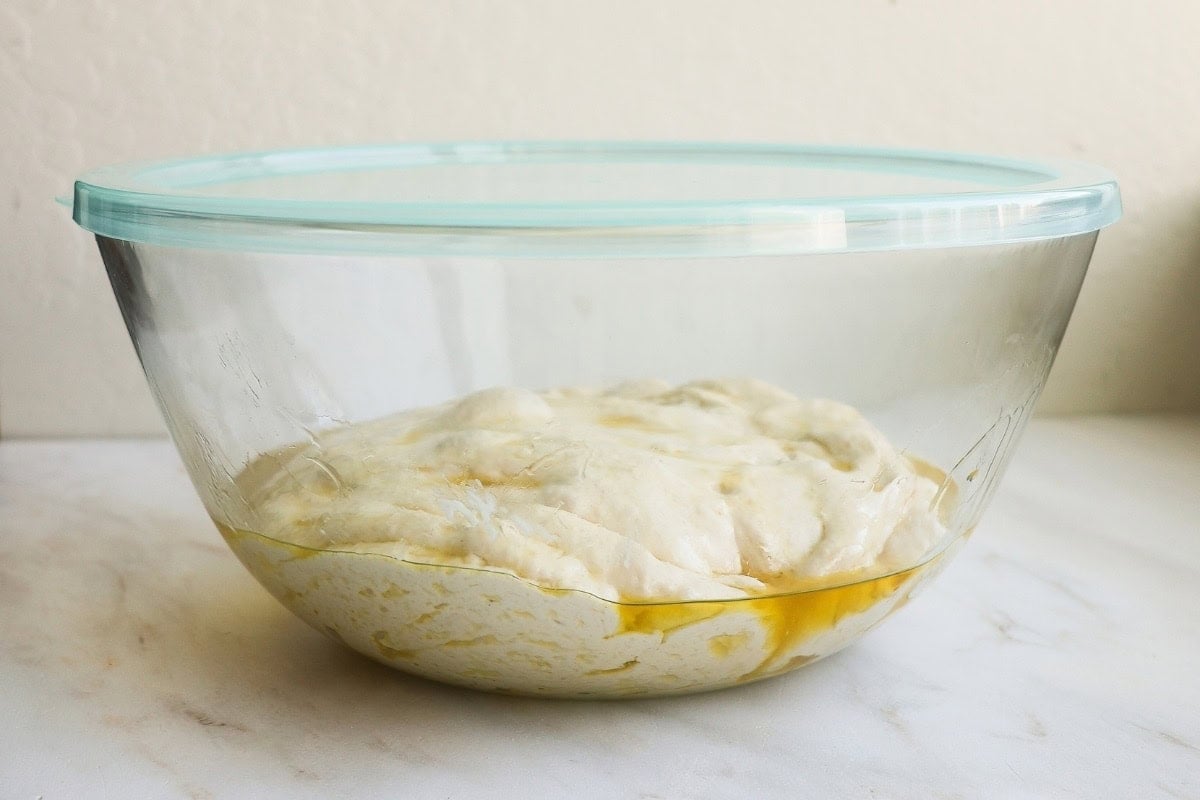 A glass bowl coated with olive oil, with focaccia dough inside and a lid on top.