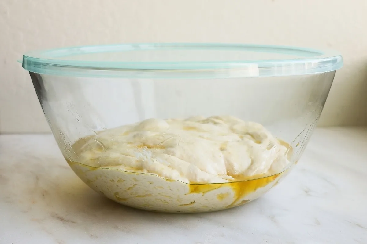 A glass bowl coated with olive oil, with focaccia dough inside and a lid on top.
