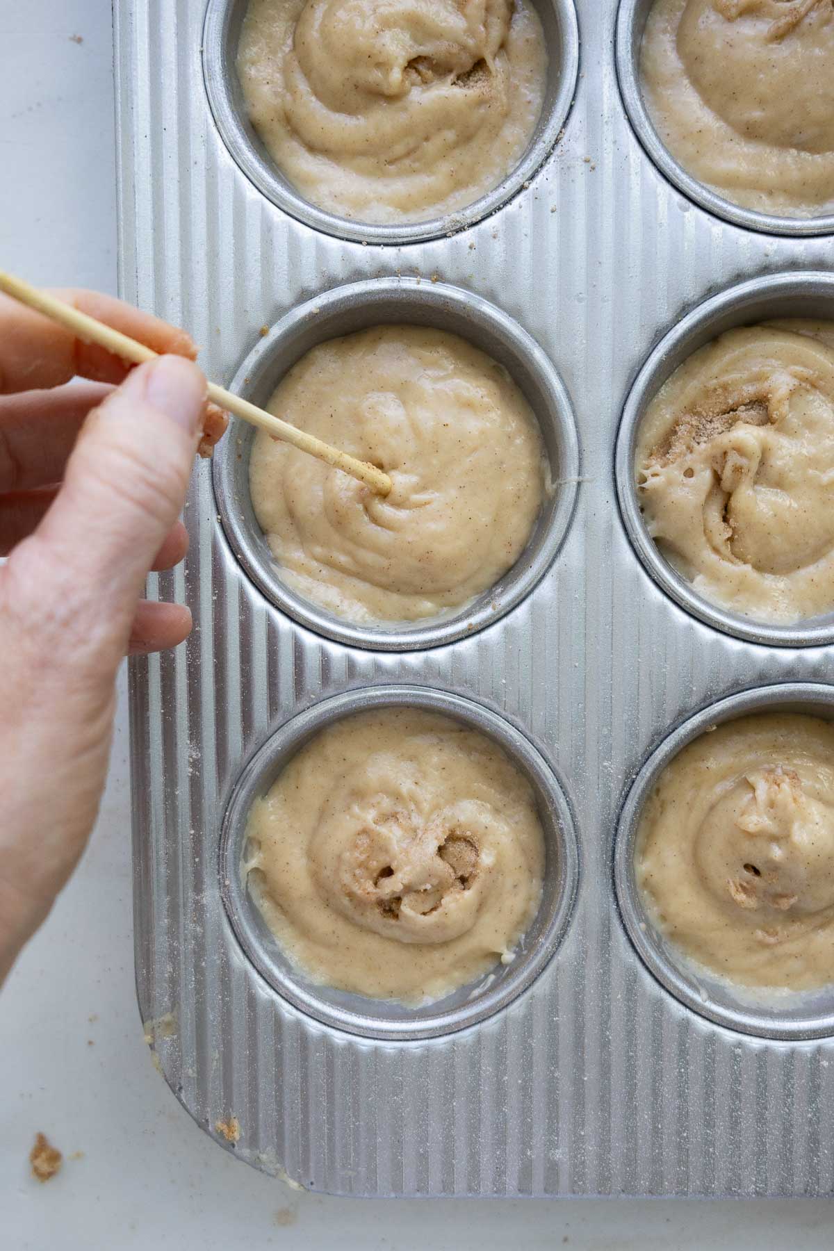 Sourdough coffee cake muffin batter in a muffin tin, with a hand holding a wooden skewer, swirling the batter in one of the muffin cups.