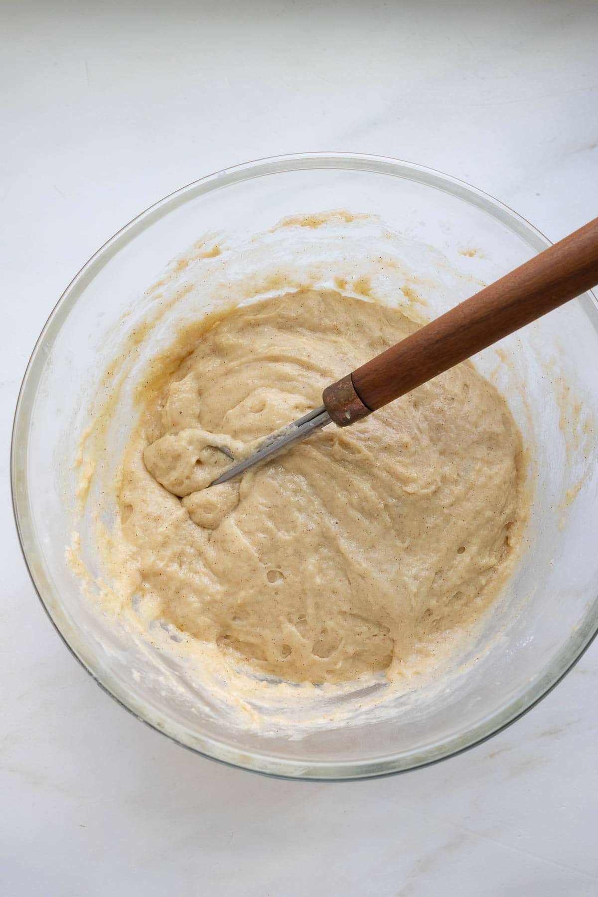 A glass bowl with sourdough coffee cake muffin batter and a Danish whisk with a wooden handle.