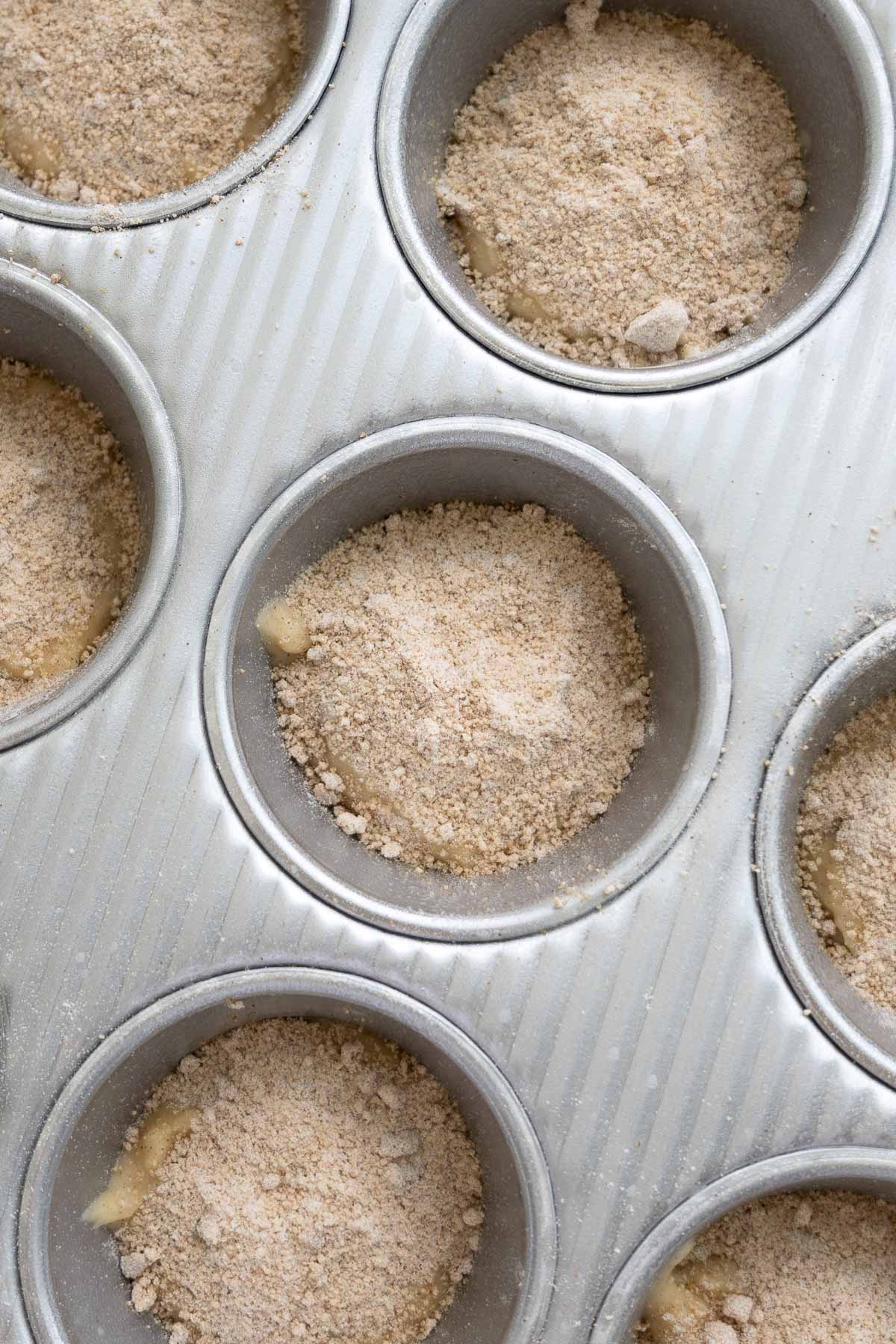 A metal muffin tin with sourdough coffee cake muffin batter and cinnamon filling on top.
