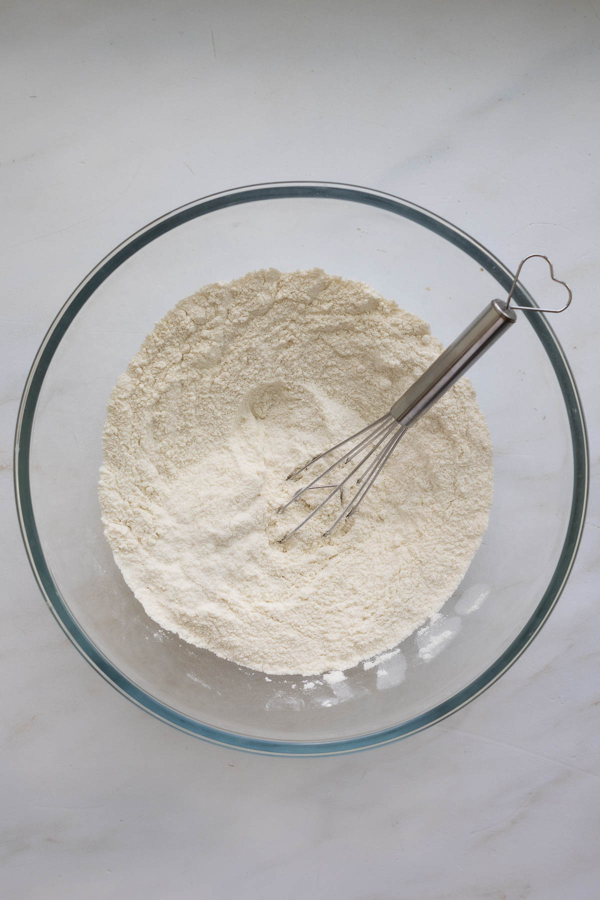 A glass bowl with a flour mixture and a wire whisk with a heart shaped handle.