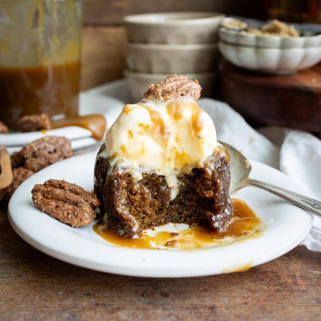 Sticky toffee pudding with a scoop of vanilla ice cream, garnished with candied pecans, on a dessert plate with dishes on the counter behind it.
